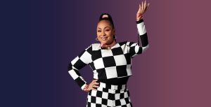 In a promotional photo for Raven’s Home, Raven-Symoné wears a black and white checkered sweater and matching pants. She is wearing a silk pink shirt and a matching pink silk ribbon in her hair. She is accessorized with gold hoop earrings and rings. She is smiling and posing with one hand on her hip and the other bent upward.