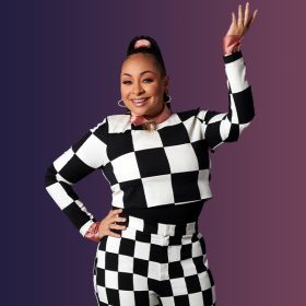 In a promotional photo for Raven’s Home, Raven-Symoné wears a black and white checkered sweater and matching pants. She is wearing a silk pink shirt and a matching pink silk ribbon in her hair. She is accessorized with gold hoop earrings and rings. She is smiling and posing with one hand on her hip and the other bent upward.