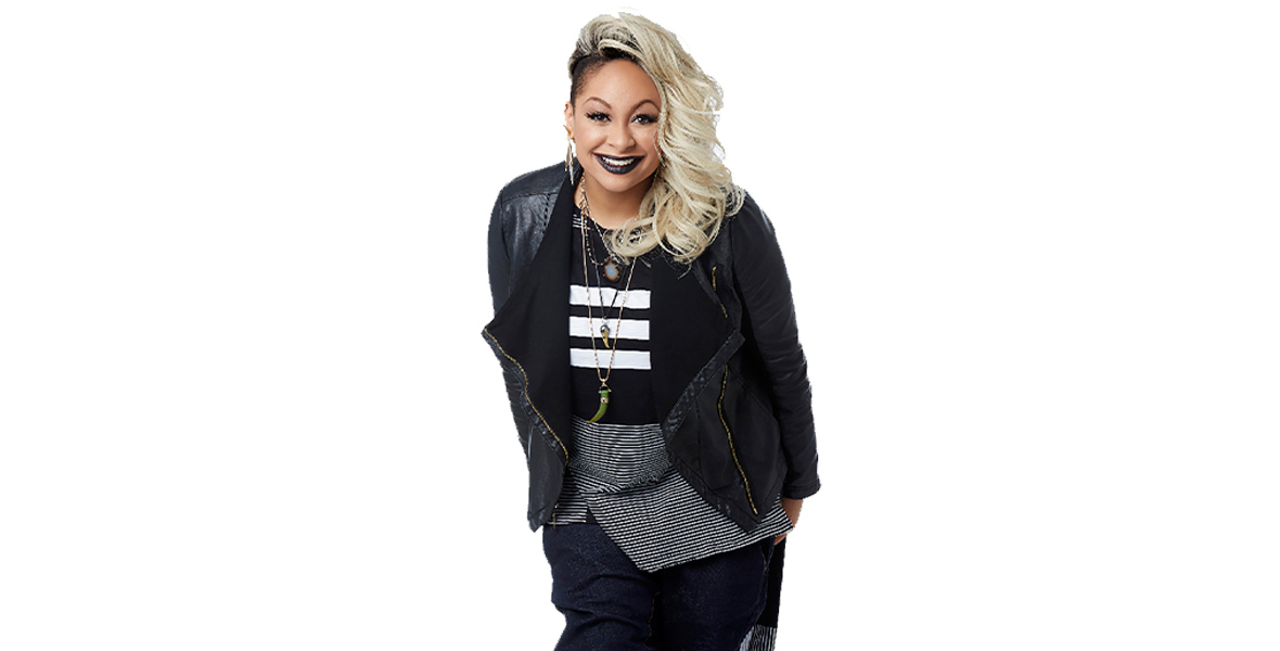 Both of Raven-Symoné’s hands are by her sides and she is leaning forward. She is wearing graphic shirt, which has thinner black and white stripes on the bottom and thicker black and white stripes on top, as well as a black leather jacket, dark jeans, layered necklaces, and gold feather earrings. She is wearing black lipstick and smiling. Her blond hair is curly and cascades to one side; the other side of her head is buzzed.