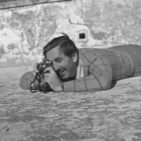 A black and white photo of Walt Disney lying flat on his stomach outside on the ground. He presses his eye to a small camera. In the corner, part of a bike’s wheel is visible.