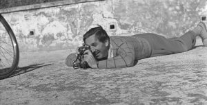 A black and white photo of Walt Disney lying flat on his stomach outside on the ground. He presses his eye to a small camera. In the corner, part of a bike’s wheel is visible.
