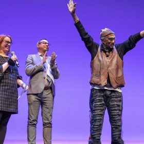 On the stage, Garth Fagan stands with his arms in the air as he accepts applause from the audience and those standing with him. Sara Nash and Greg Reiner stand beside him, smiling and clapping, as do three others pictured. The stage’s backdrop is bright purple.