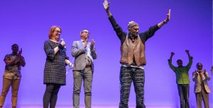 On the stage, Garth Fagan stands with his arms in the air as he accepts applause from the audience and those standing with him. Sara Nash and Greg Reiner stand beside him, smiling and clapping, as do three others pictured. The stage’s backdrop is bright purple.