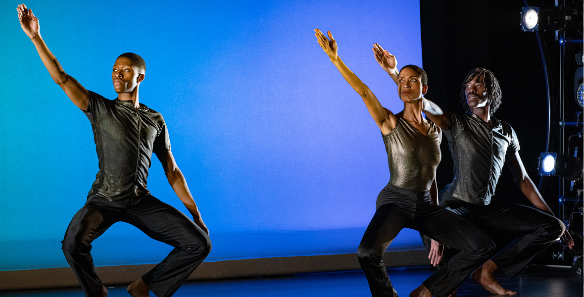 Three dancers perform on stage, crouching on their toes and raising their right arm diagonally toward the ceiling. They wear simple silver shirts and black pants. The backdrop is bright blue.