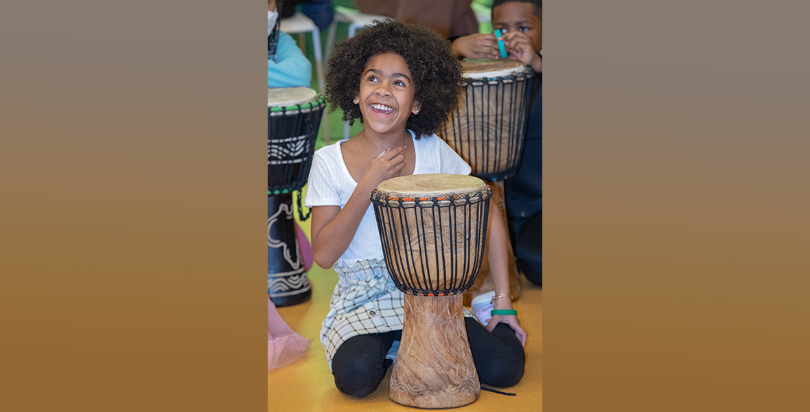 A little girl kneels in front of a drum, grinning off to her right. She wears a white shirt, a plaid skirt, and black tights.