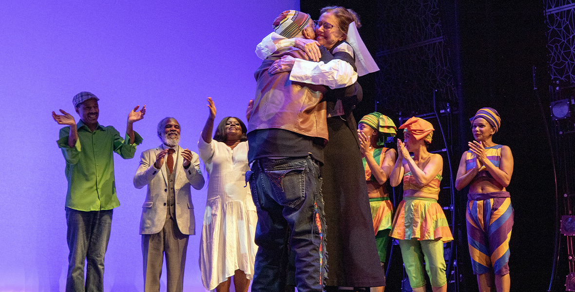 On stage, Garth Fagan and Julie Taymor hug each other as performers smile and clap behind them. The backdrop is bright purple.