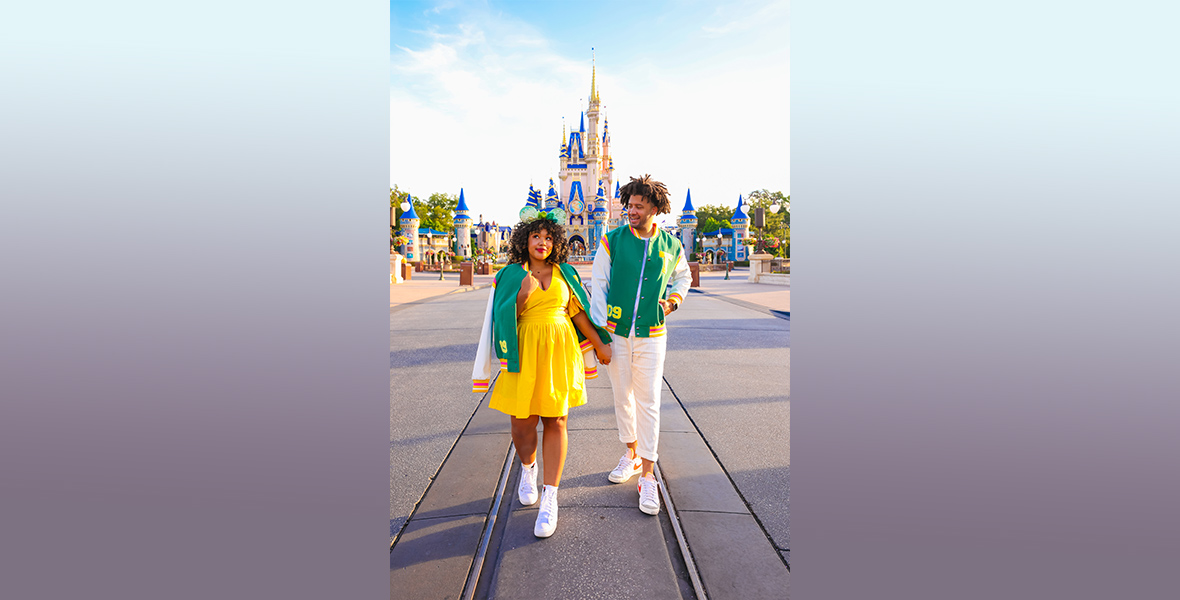 Courtney Quinn and her fiancé, Paris, hold hands while walking down Main Street, U.S.A at Magic Kingdom, Cinderella Castle shining brightly in the sun behind them. Courtney is wearing a yellow dress, Tiana ears, and the Princess Tiana collection letterman jacket draped loosely around her shoulders. One hand is outstretched behind her, holding onto Paris, who is wearing the same letterman jacket and white pants.