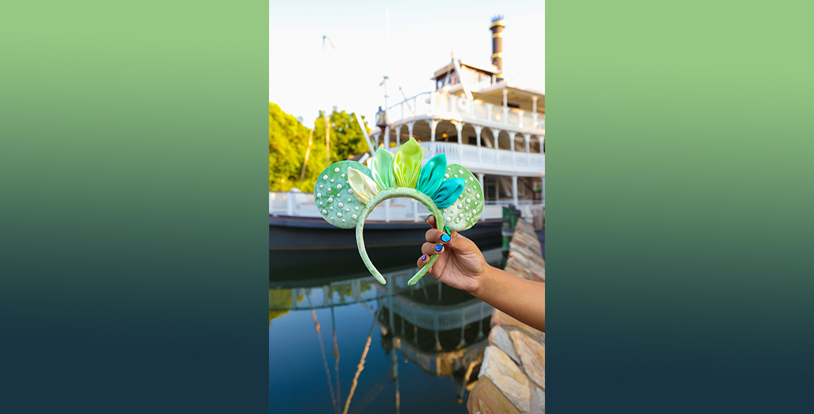 A solo image of the Princess Tiana mouse ears. Courtney Quinn’s hand is holding the ears in front of a blurry Mark Twain Riverboat at Disneyland. The ears are a mint green, velvet material with pearl studs all over the ears. Across the headband and white, green, and blue petals.