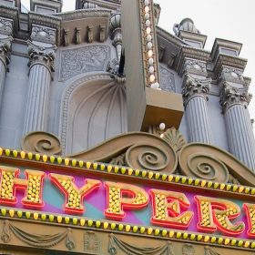 An exterior shot of the Hyperion Theater at Disney California Adventure Park. It is daytime and the top of the marquee is visible, showcasing large, capitalized, serif letters spelling out "Hyperion" in yellow light bulbs surrounded by a red trim. Towering above the marquee are grey, stone columns dotted with blinking white lights. A painted city background is barely visible to the left of the theater entrance.