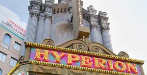 An exterior shot of the Hyperion Theater at Disney California Adventure Park. It is daytime and the top of the marquee is visible, showcasing large, capitalized, serif letters spelling out "Hyperion" in yellow light bulbs surrounded by a red trim. Towering above the marquee are grey, stone columns dotted with blinking white lights. A painted city background is barely visible to the left of the theater entrance.