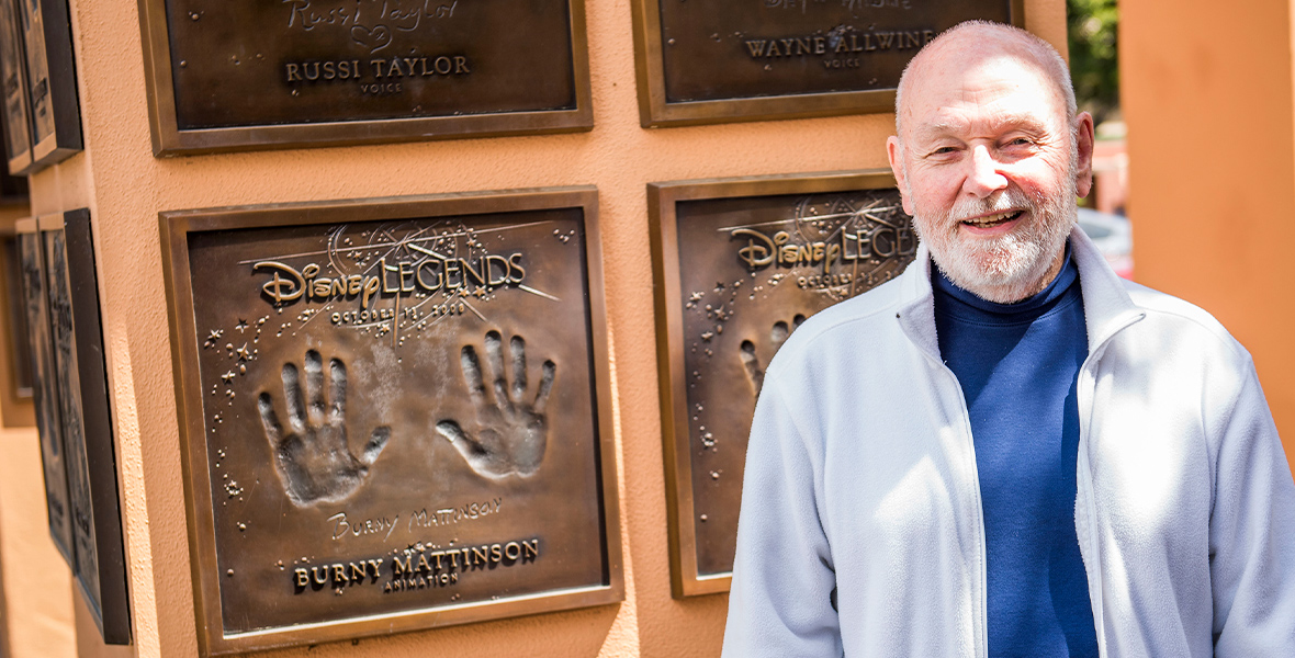 Disney Legend Burny Mattinson stands in front his bronzed handprints and bronzed signature at Legends Plaza. He is smiling and wearing a cerulean turtleneck sweater and an unzipped, powder blue fleece jacket. Mattinson has a full, white beard.