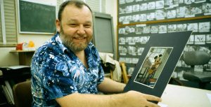 Disney Legend Burny Mattinson sits at an animator’s desk and holds up a Mickey Mouse cel. He is smiling and wearing a blue tropical button-up shirt. Behind him are black and white storyboards, and on his desk is a sketch of Ebenezer Scrooge.