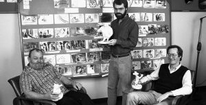 Disney Legend Burny Mattinson reclines in a leather chair; Ron Clements stands to his left, and John Musker sits in a leather chair to his far left. Each holds a white maquette from The Great Mouse Detective. Behind them are film storyboards.