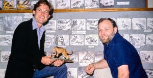 Disney Legend Kurt Russell kneels as he holds a maquette for The Fox & The Hound. To his left is Disney Legend Burny Mattinson, who is also kneeling. Russell is wearing blue jeans, a blue and white gingham shirt, a black blazer, and tan cowboy boots. Mattinson is wearing a blue polo, khaki pants, black socks, and brown dress shoes. Behind both men are colored and black and white storyboards for the movie.