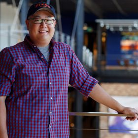 Peter Sohn smiles as he smiles for a photo just above the Pixar Animation Studios lobby, where he is resting his left hand on a railing. He is wearing an untucked blue and red checkered shirt over a gray T-shirt. He is wearing blue jeans, black rimmed glasses, and navy blue hat that says “PIXAR” in capital orange letters.
