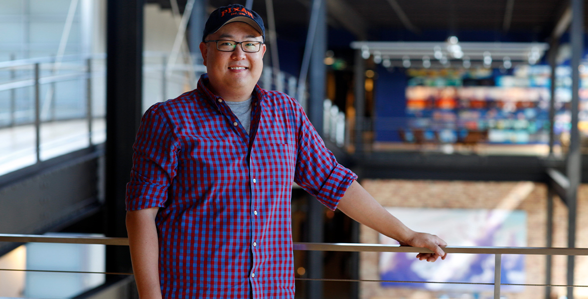 Peter Sohn smiles as he smiles for a photo just above the Pixar Animation Studios lobby, where he is resting his left hand on a railing. He is wearing an untucked blue and red checkered shirt over a gray T-shirt. He is wearing blue jeans, black rimmed glasses, and navy blue hat that says “PIXAR” in capital orange letters.
