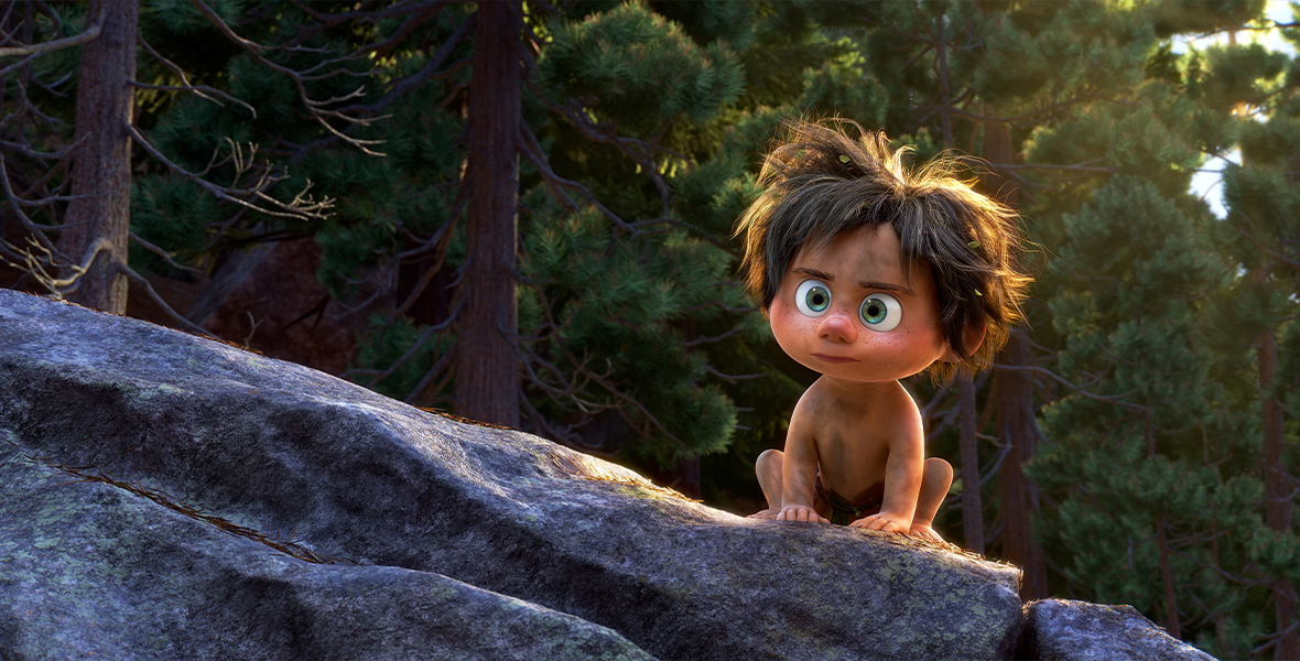 Spot, a 7-year-old caveboy, crouches on a rock. Behind him are pine trees.