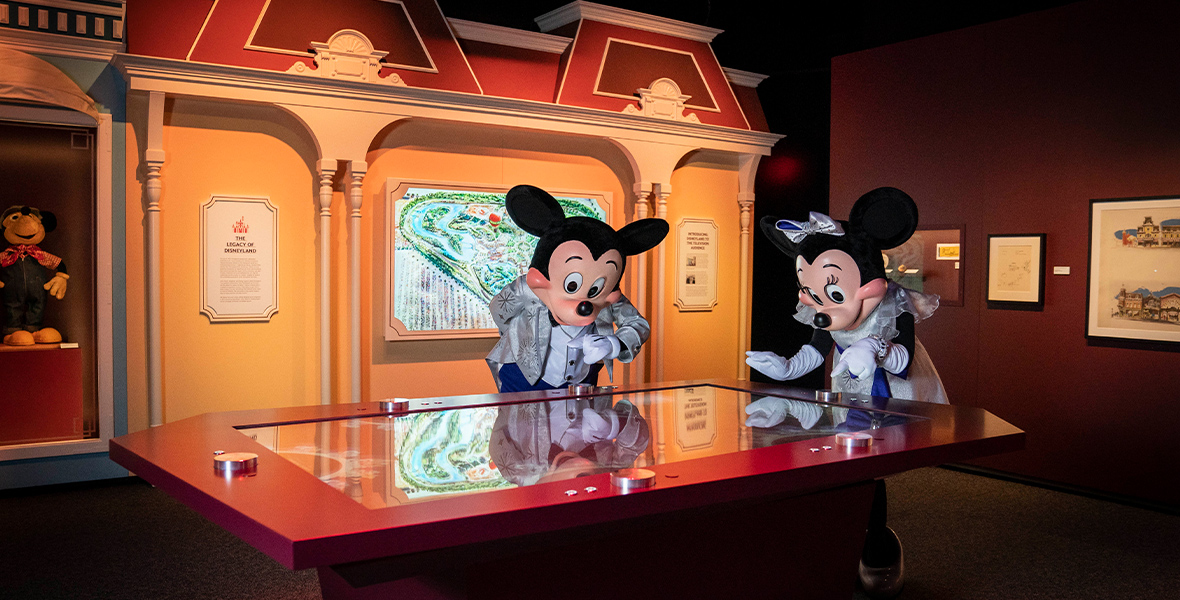 Mickey and Minnie lean over an interactive table in the “Your Disney World” gallery. Behind them is an illuminated print of Peter Ellenshaw’s concept art for Disneyland. The wall behind them is designed to look like the building fronts of Main Street, U.S.A.