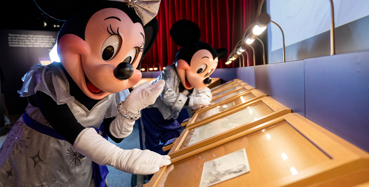 In front of a red curtain, Minnie and Mickey lean in to admire Disney artwork showcased on desks lit by small, curved lamps. Minnie holds a hand to her face in the foreground, while behind her Mickey is focused on the art in front of him.