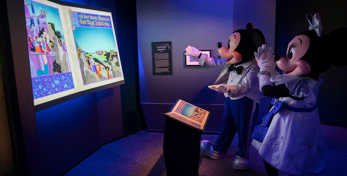 Mickey and Minnie admire a display in Disney100: The Exhibition that showcases a large-screen depiction of Eyvind Earle’s artwork inside the Sleeping Beauty prop book. Mickey holds his hand out to the image, while Minnie applauds.