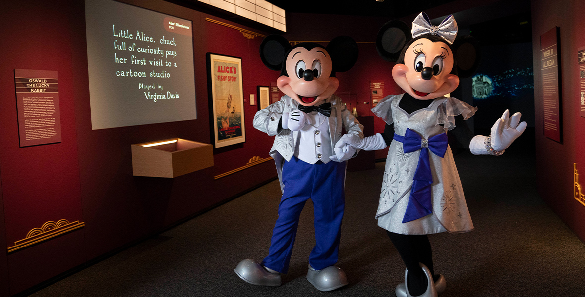 Mickey and Minnie hold hands in the “Where It All Began” gallery of Disney100: The Exhibition. They are both dressed in sparkling, platinum outfits with royal blue accents. To their left is a marquee for The Alice Comedies and a screen playing a clip from the Alice’s Wonderland short.