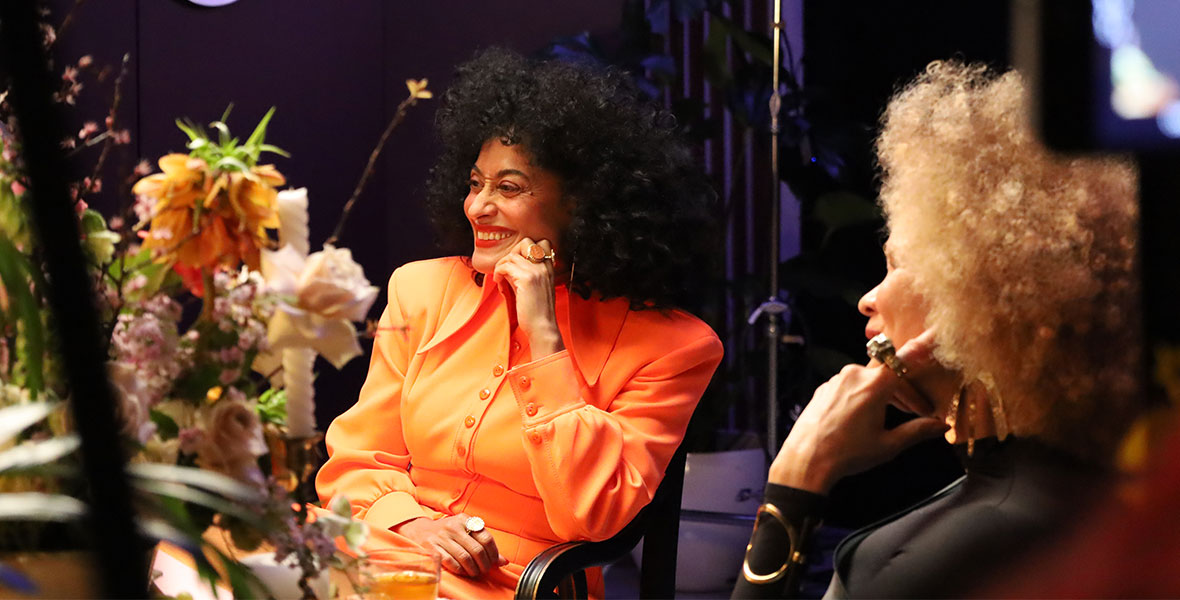 In a scene from The Hair Tales, actor and Disney Legend Tracee Ellis Ross sits in a chair next to Michaela angela Davis. Ross wears an orange blouse and smiles.