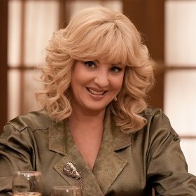 Wendi McLendon-Covey is pictured in her role as Beverly in the ABC series The Goldbergs. She has her signature mane of blond hair and is wearing a tan jacket with wide lapels. She is seated at a restaurant table with wine and water glasses in front of her.
