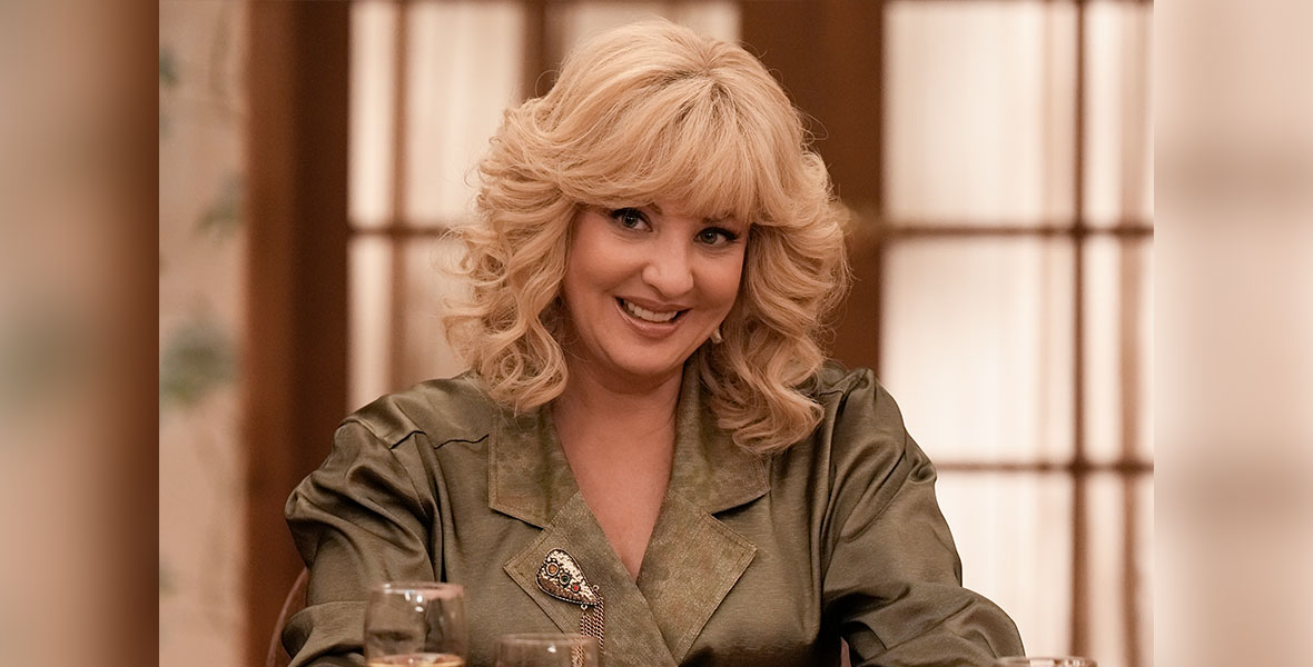 Wendi McLendon-Covey is pictured in her role as Beverly in the ABC series The Goldbergs. She has her signature mane of blond hair and is wearing a tan jacket with wide lapels. She is seated at a restaurant table with wine and water glasses in front of her.