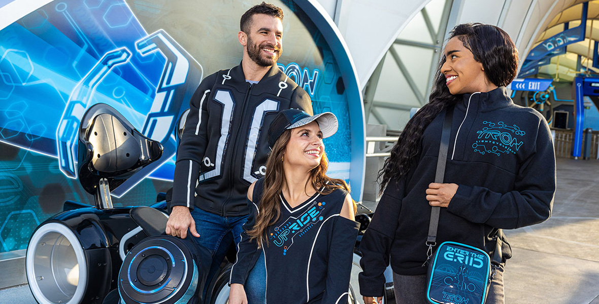 Three people wear TRON merchandise outside the attraction TRON Lightcycle / Run presented by Enterprise.