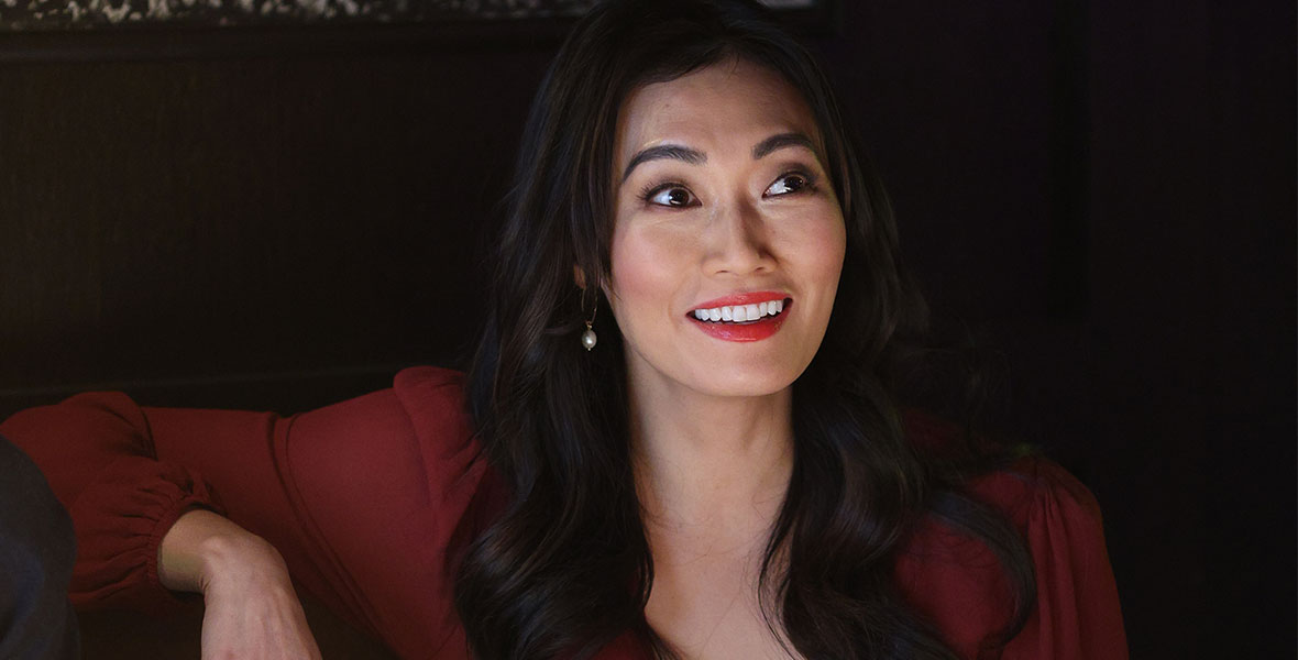 In a scene from an episode of The Company You Keep, actor Catherine Haena Kim sits in a restaurant booth and wears a red blouse.