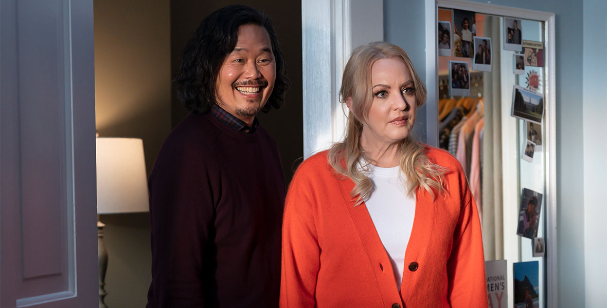 David S. Jung, on the left, and Wendy McLendon-Covey, on the right, stand in front of a doorway in a scene from the Disney Channel movie Prom Pact. The actors are portraying the parents of the main character. Jung is wearing a dark brown sweater, buttoned up high, over a plaid shirt, while McLendon-Covey has on a loose-fitting orange cardigan sweater over a white collarless shirt. Jung is smiling broadly, while McLendon-Covey looks worried.