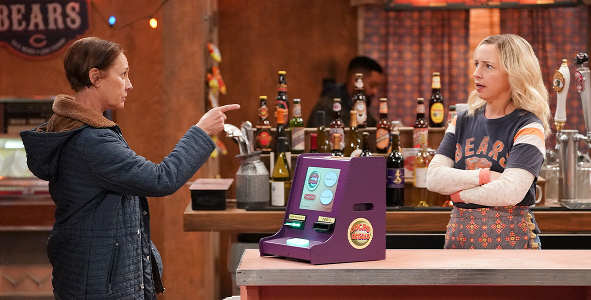 In a scene from an episode of The Conners, actor Laurie Metcalf points with her right hand at actor Alicia Goranson, who stands behind a restaurant counter. Metcalf wears a navy raincoat and denim jeans. Goranson wears a blue T-shirt with the Chicago Bears logo layered over a white long-sleeved T-shirt and a gray apron.