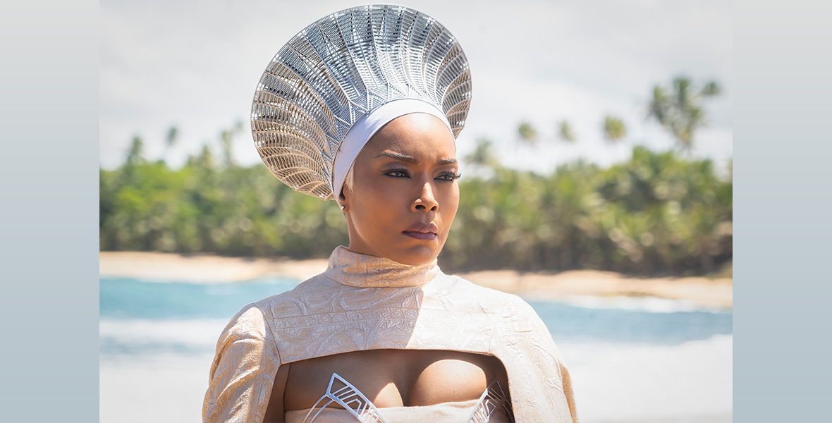 In a scene from Black Panther: Wakanda Forever, actor Angela Bassett stands on a sandy shore and wears a white tunic dress and an ornate silver and white headpiece.