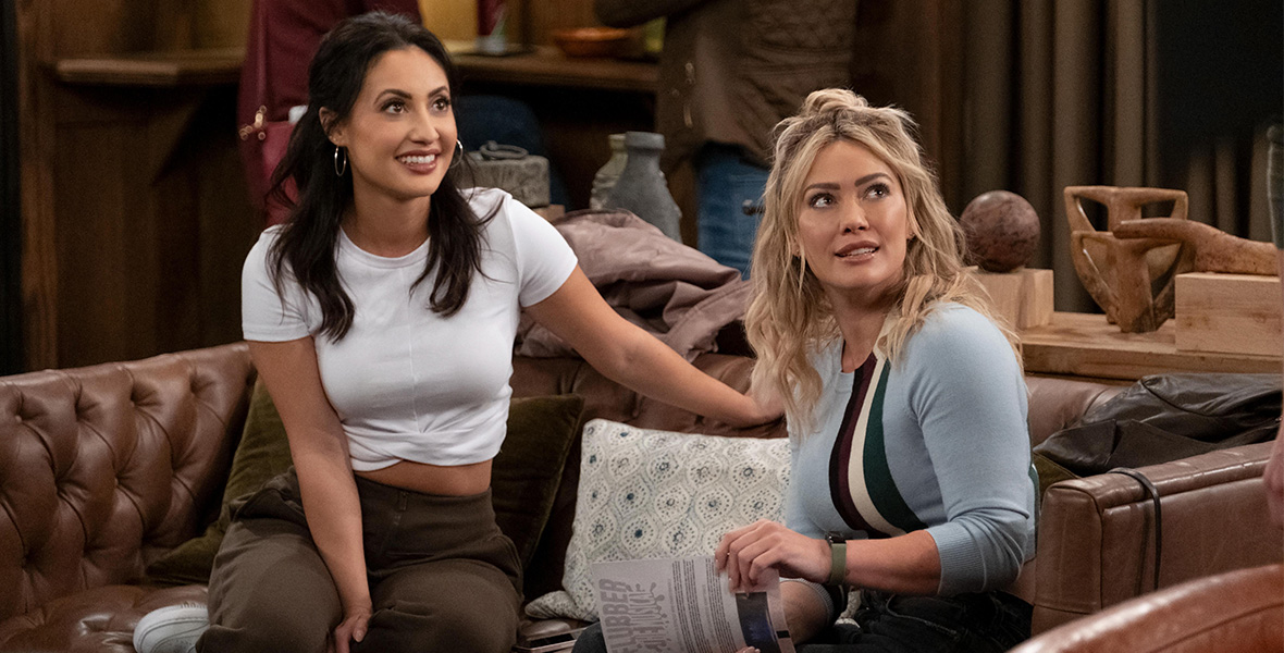 In a scene from an episode of How I Met Your Father, actors Francia Raisa and Hilary Duff sit on a brown leather couch. Raisa wears a white T-shirt and brown pants. Duff wears a light blue sweater with four vertical stripes and black jeans and holds a sheet of paper.