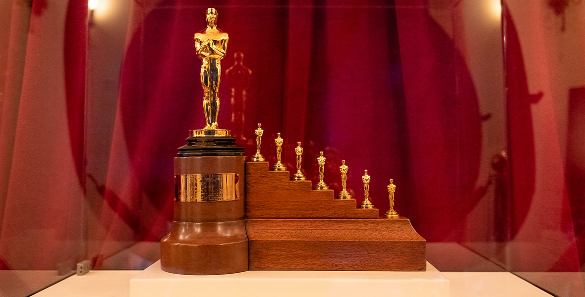 An image of the special Snow White-themed Oscar statue that Walt Disney received in 1939, as currently displayed at Carthay Circle Restaurant inside Disney California Adventure Park at Disneyland Resort. Behind the gold statue—which has one “regular” size Oscar and seven very small ones lined up on small wooden steps next to it—is a red curtain.