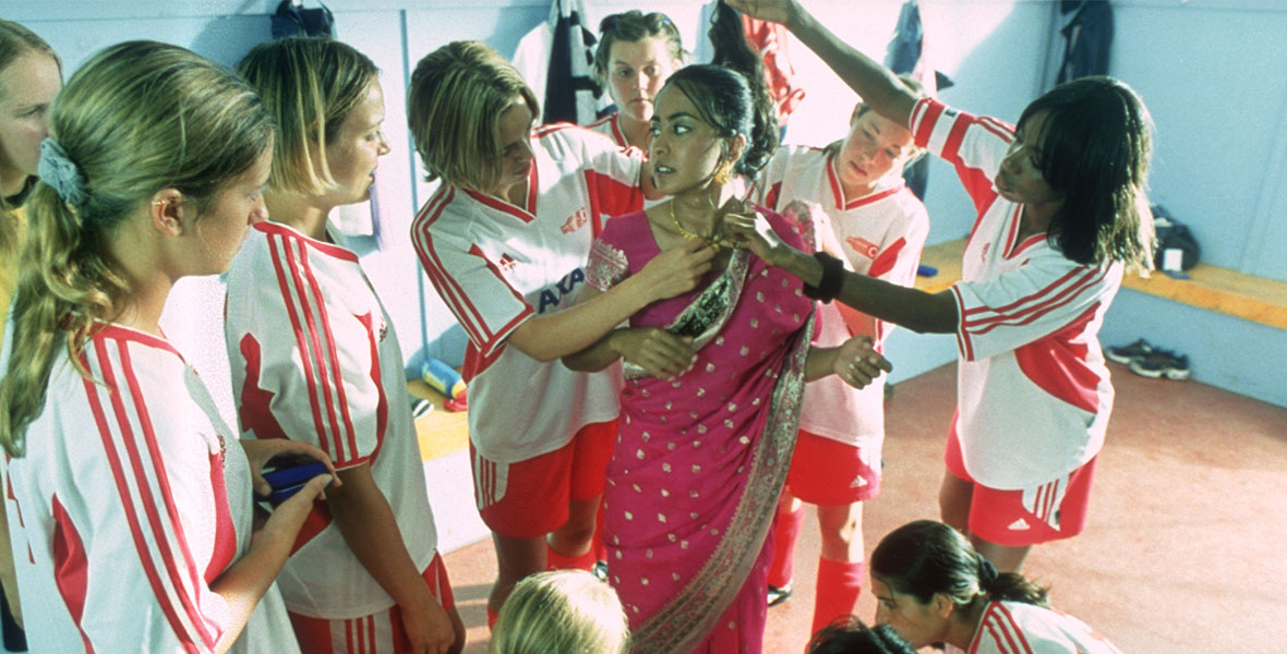 In a scene from Bend It Like Beckham, actor Parminder Nagra is surrounded by her teammates who hold her dress in a hot pink and gold sari.
