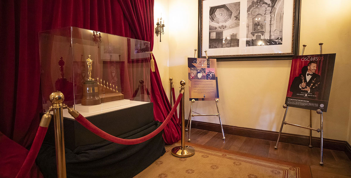 A photo of the Snow White-inspired Oscar statue display inside Carthay Circle Restaurant in Disney California Adventure Park. The statue sits in a glass display behind red velvet ropes with a red curtain backdrop. The one-of-a-kind statue consists of one regular-sized, golden statue with seven tiny statues beside it.