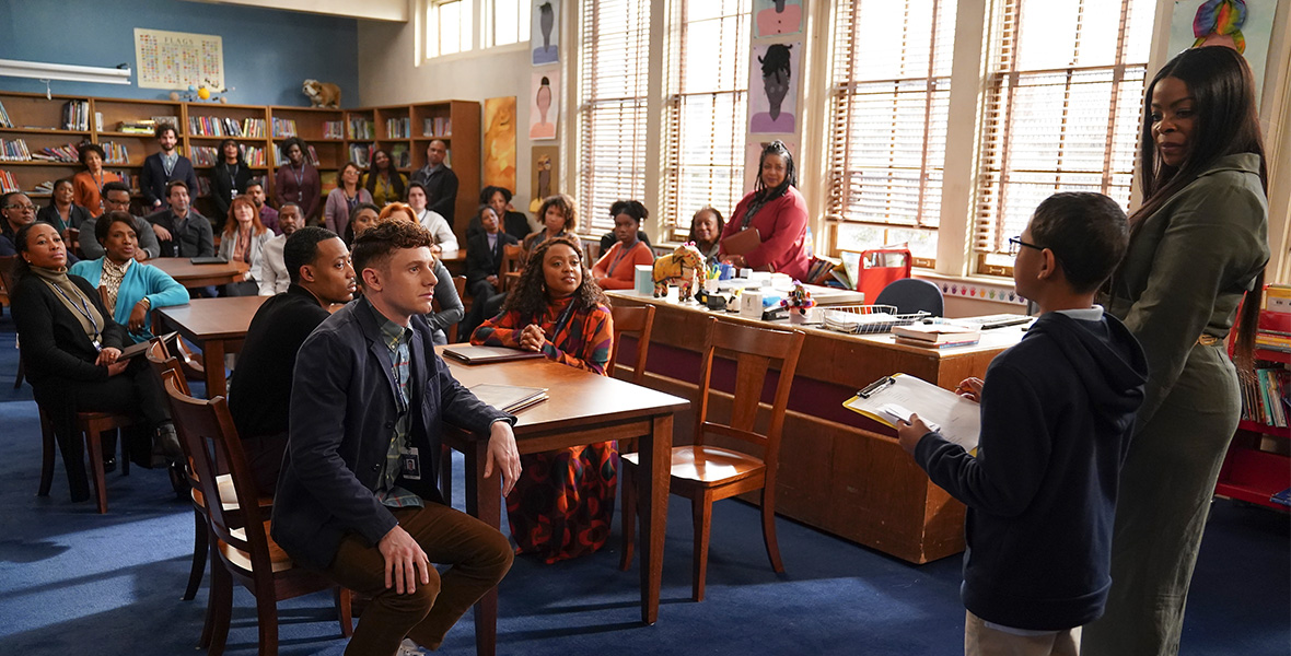 In a scene from an episode of Abbott Elementary, actor Janelle James stands with a young male student in the library and speak to the school’s teaching staff, who sit at wooden tables. 