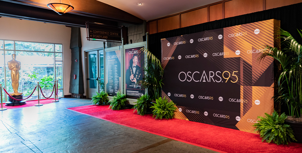 An image of the Oscars “step and repeat”-style red-carpet display at Disney’s Grand Californian Hotel & Spa. A backdrop is seen toward the center-right of the image with the “Oscars95” logo on it; toward left of the backdrop is a poster featuring Oscars host Jimmy Kimmel. To the far left is a giant Oscar statue in front of a window, surrounded by a velvet rope.