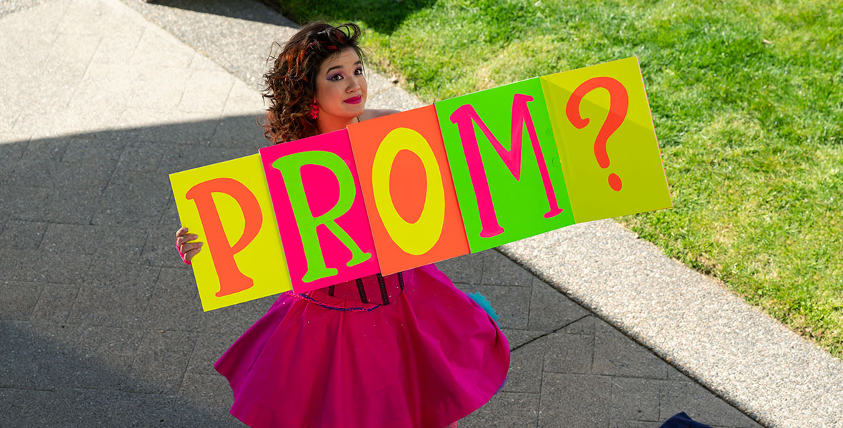 In a scene from Prom Pact, actor Peyton Elizabeth Lee holds a neon sign reading, “Prom?” and wears a hot pink dress, hot pink tights, and hot pink shoes.