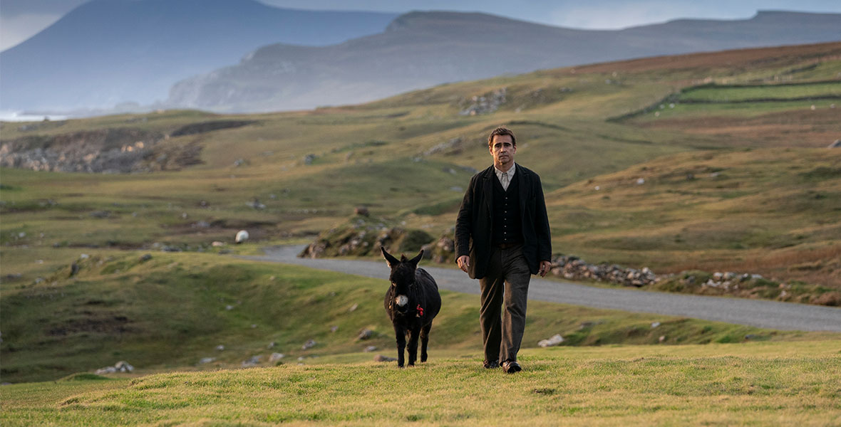 In a scene from the film The Banshees of Inisherin, Colin Farrell as Pádraic Súille walks across a vast expanse of rolling green hills. A small black donkey trots beside him.
