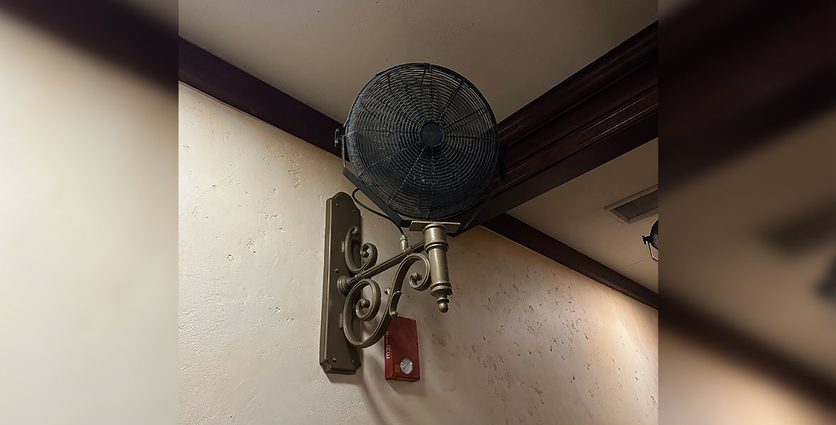 A black metal fan is mounted on a beige concrete wall. The fan is attached to a bronze wall mount with ornate details. Wooden beams can be seen extending across the ceiling of the room and a fire alarm sits behind the fan on the wall.