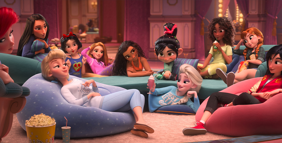 In a scene from Ralph Breaks the Internet, Vanellope von Schweetz, a young female character, sits center in a large room and surrounded by Disney Princesses—Ariel, Pocahontas, Cinderella, Snow White, Rapunzel, Moana, Elsa, Tiana, Anna, and Mulan—who are dressed in contemporary, casual clothing.