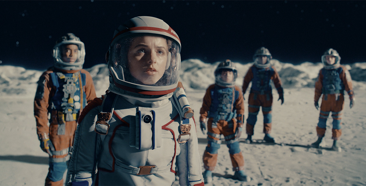 In an image from Disney+’s Crater, Mckenna Grace as Addison (as well as several other cast members to her left and right) are wearing space suits and are standing on the lunar surface, staring up at something off camera and to the right.