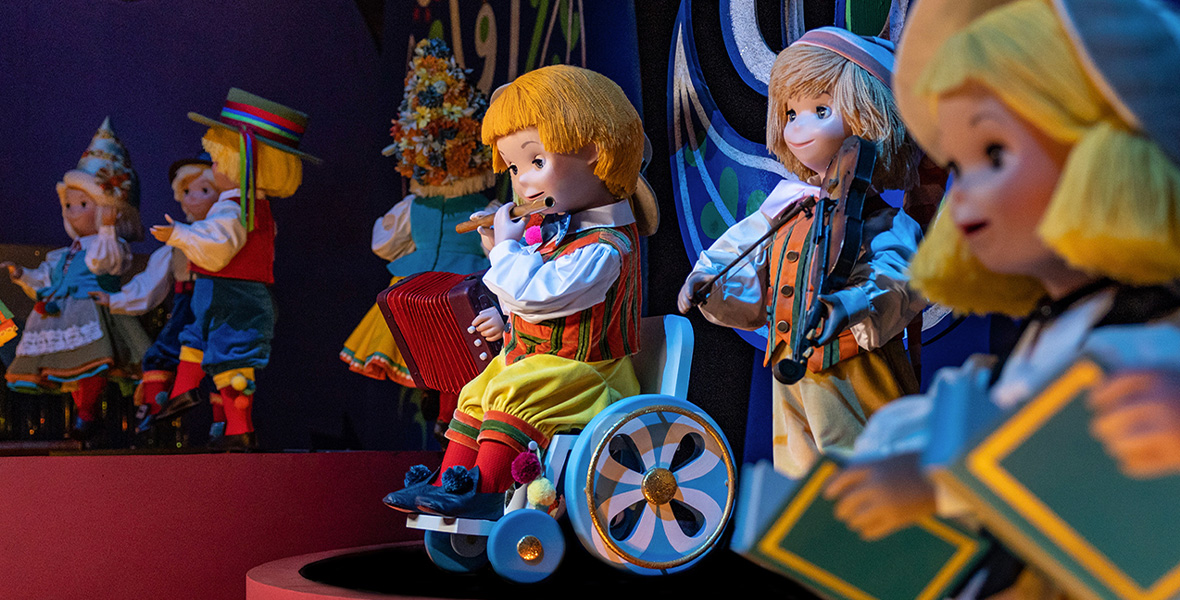 In a new image from the it’s a small world attraction at Magic Kingdom Park at Walt Disney World Resort, a new doll can be seen sitting in a wheelchair. The doll is playing a flute, has blonde hair, and is wearing a white shirt, an orange and green striped vest, and yellow pants. Around the doll are other it’s a small world dolls dressed in their typical international-style outfits.
