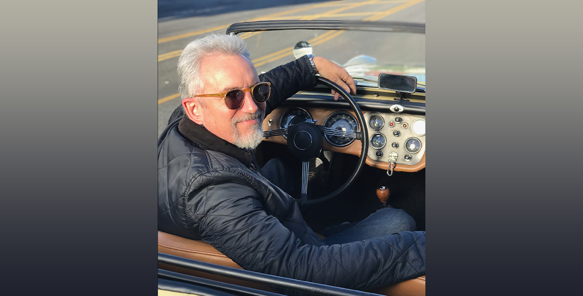 A headshot of Jeff “Swampy” Marsh, of Phineas and Ferb fame. He’s behind the wheel of a classic convertible car, with his left arm resting on the steering wheel. He has gray hair and is wearing sunglasses and a puffy black jacket.