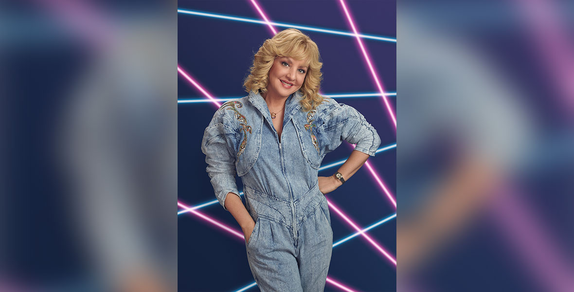 Wendi McLendon-Covey, in character as Beverly from The Goldbergs, poses in a denim jumpsuit in front of a black background criss-crossed by glowing pink and blue lines. She is smiling, with one hand on her hip and one in a lower pocket.