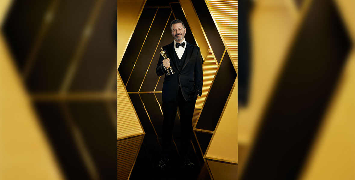 A poster of Jimmy Kimmel, who wears a black suit and bow tie. He clutches an Oscar in his right hand and smiles at the camera, his other hand is tucked into his pocket. The background is bright black and gold.