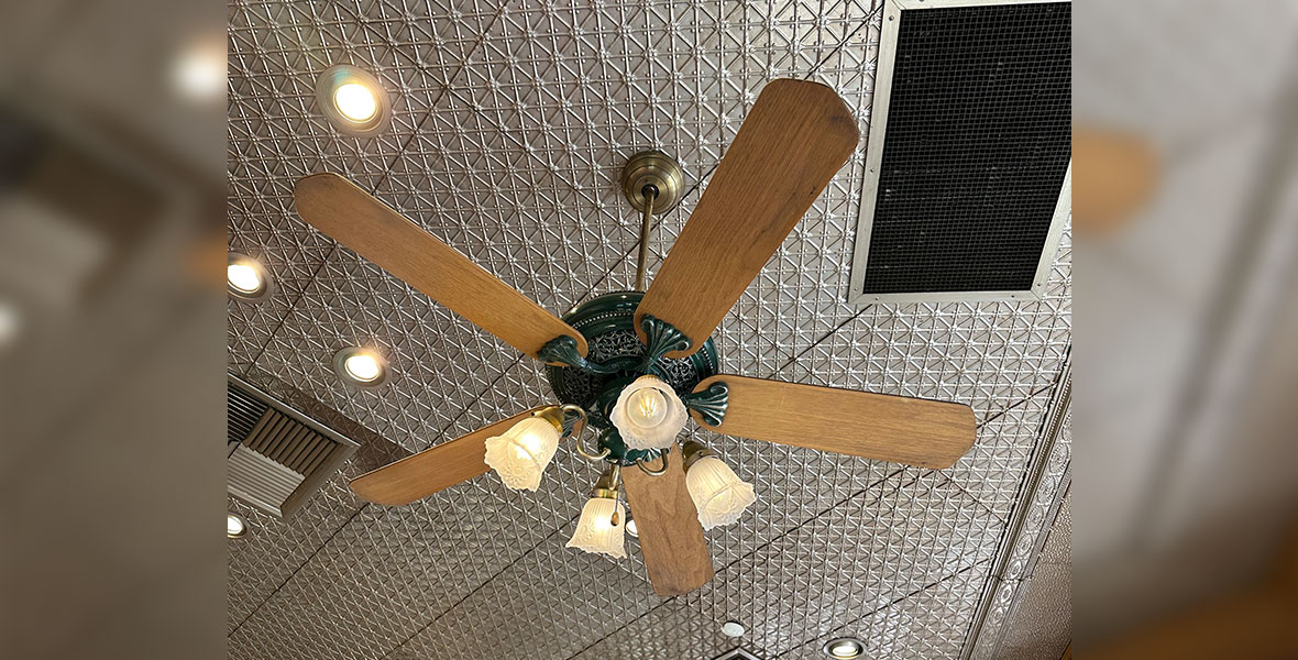 A ceiling fan with wooden blades, black metal details, and white glass lightbulb holders is hanging from a checker marked silver colored ceiling.