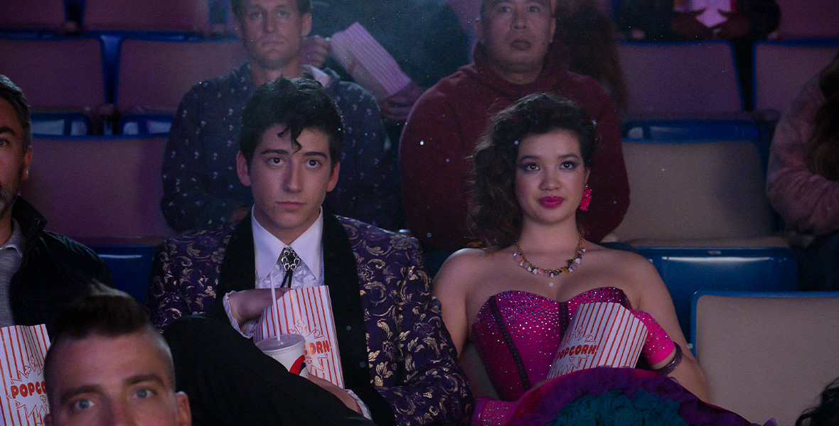 In a scene from Prom Pact, actors Milo Manheim and Peyton Elizabeth Lee sit in a movie theater and eat popcorn. Manheim wears a white button-down shirt, a purple pattered suit jacket. And a black bolo tie. Lee wears a hot pink, strapless, sequin dress and matching gloves.